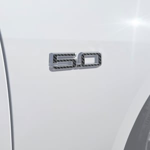 May include: A close-up of a white car with a black and silver "5.0" emblem on the side. The emblem has a carbon fiber pattern.