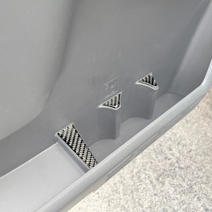 May include: Close-up of a gray car interior door panel with three carbon fiber trim pieces. The trim pieces are in the shape of a triangle and are located in the door panel's storage compartment.