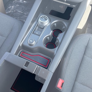 Puede incluir: Un interior de coche negro con un ribete rojo alrededor de la consola central. La consola tiene un portavasos, una palanca de cambios y un compartimento de almacenamiento. La consola está cubierta con una alfombra negra con un ribete rojo.