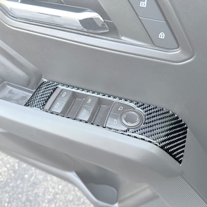 Può includere: Primo piano di un pannello porta interno di un'auto con un kit di rivestimento in fibra di carbonio. Il kit di rivestimento copre i comandi dei finestrini e include un pulsante per la serratura della porta.