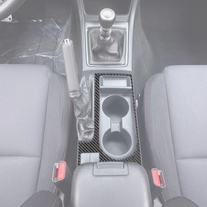 Puede incluir: Un kit de moldura de consola central de fibra de carbono negro para un automóvil. El kit de moldura cubre el área alrededor de los portavasos y la palanca de cambios.