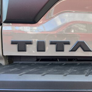 May include: A close-up of a gray truck bed with a black carbon fiber "TITAN" emblem.
