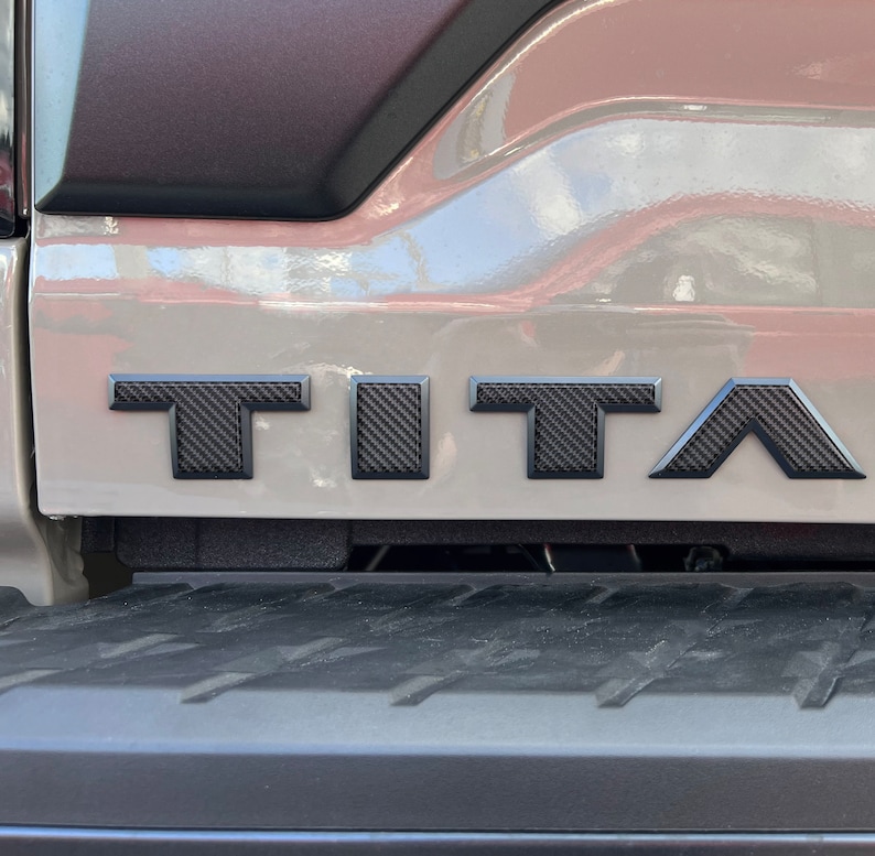 May include: A close-up of a gray truck bed with a black carbon fiber "TITAN" emblem.