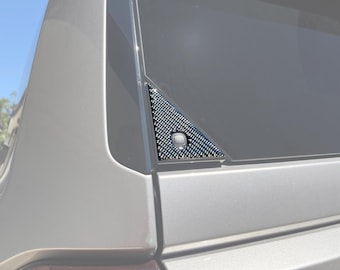 May include: A close-up of a silver car with a black carbon fiber triangle trim piece on the rear window. The trim piece has a small button in the center.