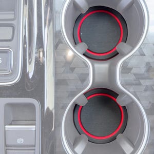 May include: A close-up of a car's interior, featuring a dual cup holder with a gray finish and red accent rings. The console also includes a parking brake button and gear shift controls.