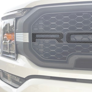 Puede incluir: Una camioneta Ford F-150 blanca con una inserción de parrilla de fibra de carbono negra. La inserción de la parrilla tiene un patrón de panal y la palabra "FORD" es visible en el centro.