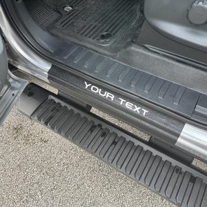 May include: Close-up of a black car door sill with a white "YOUR TEXT" inscription. The sill has a textured surface and is designed to protect the car's paint.