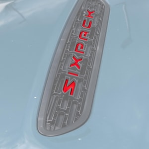 May include: A gray automotive hood vent featuring a black grille insert and the word SIXPACK displayed in bold, bright red lettering. The vent is integrated into a smooth, light gray painted surface, showcasing a clean and modern design.