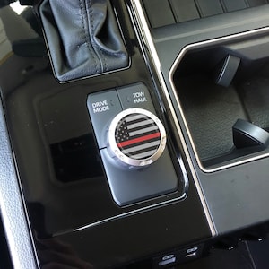 May include: A black and silver car knob with a thin red line and an American flag design. The knob is labelled "Drive Mode" and "Tow/Haul".