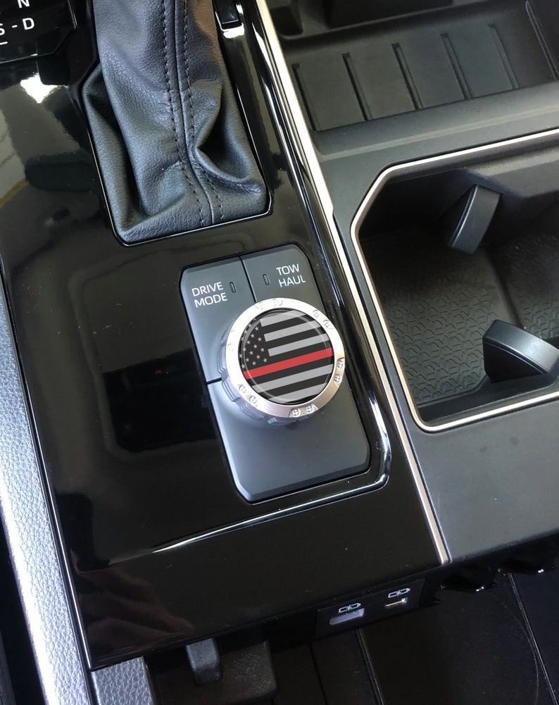 May include: A black and silver car knob with a thin red line and an American flag design. The knob is labelled "Drive Mode" and "Tow/Haul".