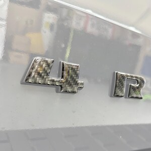 Puede incluir: Primer plano de un emblema de coche con los números "4" y las letras "R U". El emblema tiene un patrón de fibra de carbono y está sobre un fondo gris claro.