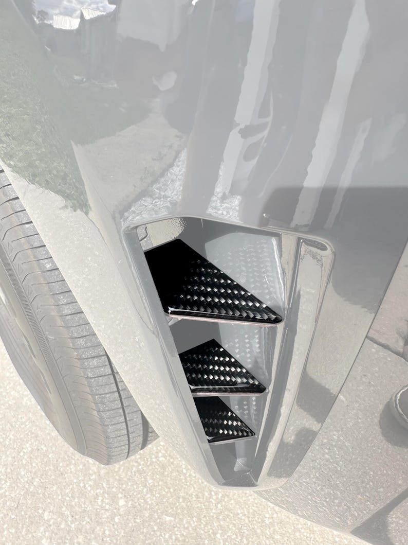 Puede incluir: Primer plano del parachoques delantero de un coche con tres cubiertas de ventilaci&oacute;n triangulares negras. Las cubiertas tienen un patr&oacute;n de tejido de fibra de carbono, lo que a&ntilde;ade una est&eacute;tica deportiva. La carrocer&iacute;a del coche es gris claro.