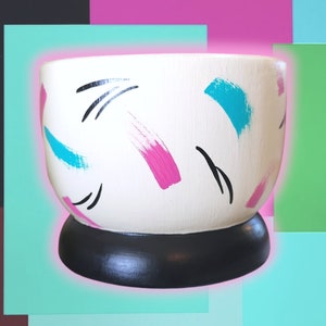 May include: A white ceramic planter with a black base, adorned with strokes of blue, pink, and black paint. The planter has a rounded shape and is suitable for indoor plants. The design is modern and colorful.