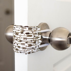 May include: A white door with a silver door knob and a brown and white patterned door stop. The door stop is a fabric cover with a pattern of arrows and hearts.