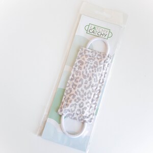 Puede incluir: Una mascarilla facial de estampado de leopardo blanco y gris con el&aacute;sticos de oreja blancos. La mascarilla est&aacute; envasada en una bolsa de pl&aacute;stico transparente con una etiqueta blanca y verde que dice "Latchy Catchy".