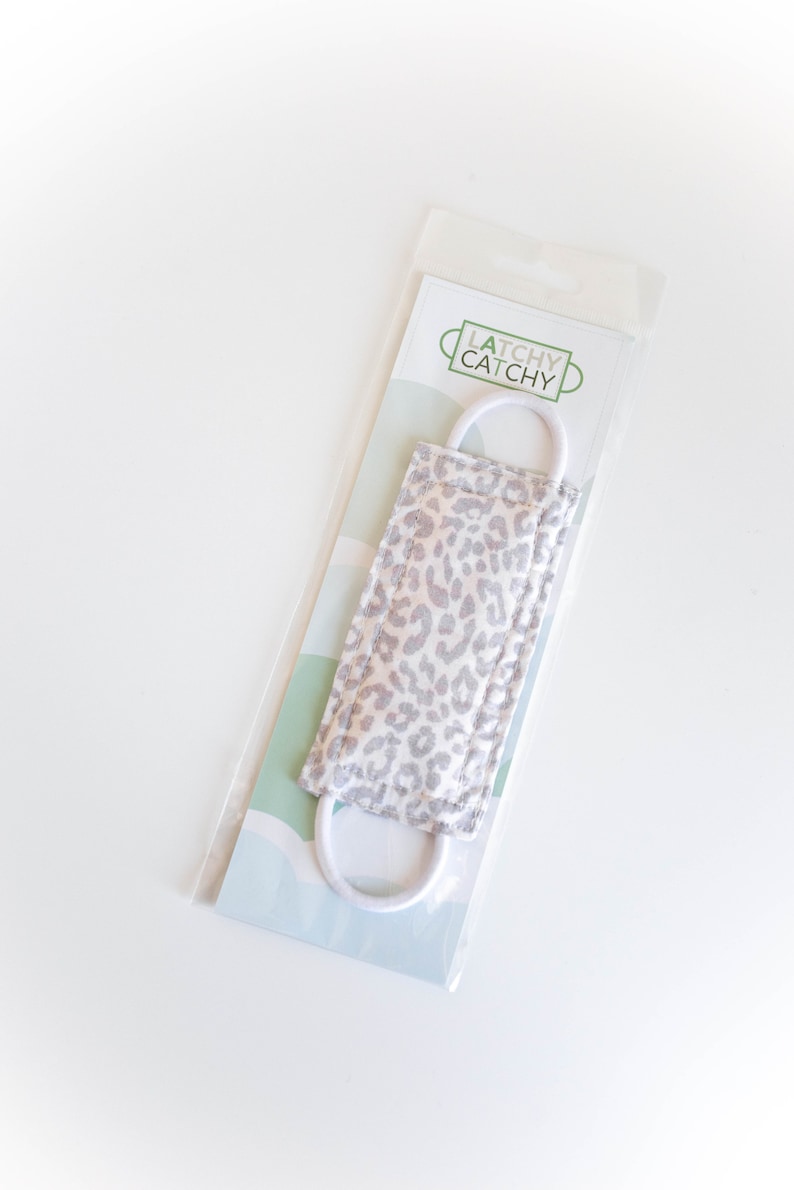 Puede incluir: Una mascarilla facial de estampado de leopardo blanco y gris con el&aacute;sticos de oreja blancos. La mascarilla est&aacute; envasada en una bolsa de pl&aacute;stico transparente con una etiqueta blanca y verde que dice "Latchy Catchy".