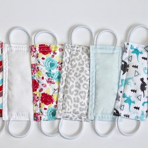 Puede incluir: Siete mascarillas de tela para la cara con lazos el&aacute;sticos blancos para las orejas. Las mascarillas presentan diferentes patrones, incluyendo un chevron rojo y blanco, un patr&oacute;n floral blanco, un estampado de leopardo gris, un color s&oacute;lido azul claro, un patr&oacute;n blanco con dinosaurios negros y un color s&oacute;lido rosa.