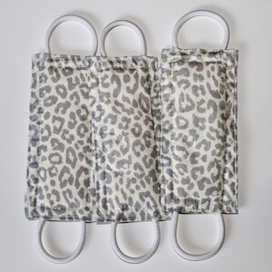 Puede incluir: Tres mascarillas de tela con estampado de leopardo gris y blanco con el&aacute;sticos de oreja blancos.