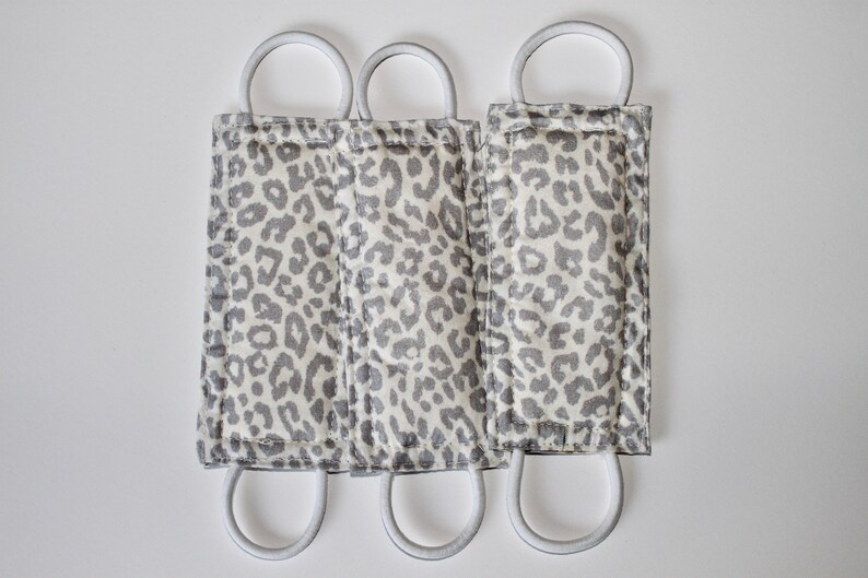 Puede incluir: Tres mascarillas de tela con estampado de leopardo gris y blanco con el&aacute;sticos de oreja blancos.