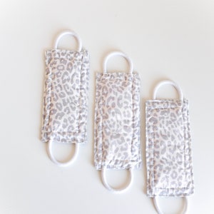 Puede incluir: Tres mascarillas de tela blanca con un estampado de leopardo gris y el&aacute;sticos de oreja blancos.