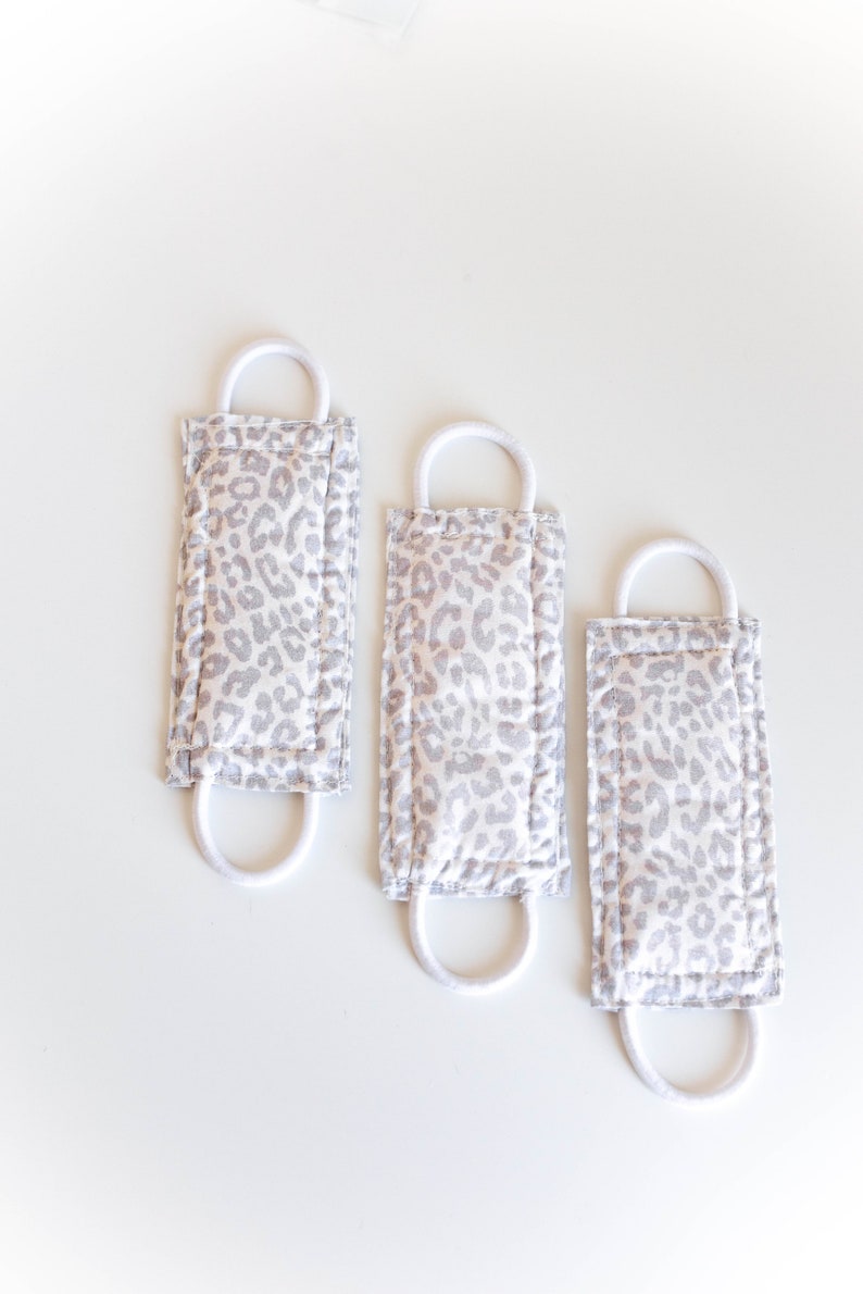 Puede incluir: Tres mascarillas de tela blanca con un estampado de leopardo gris y el&aacute;sticos de oreja blancos.