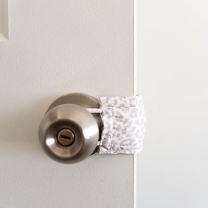 Puede incluir: Una puerta blanca con una perilla de puerta plateada y un tope de puerta de tela blanca y gris con estampado.