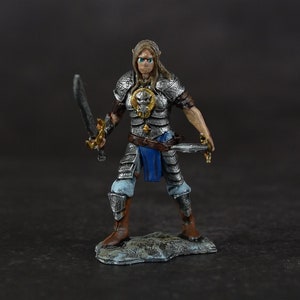Puede incluir: Una figura en miniatura de una guerrera que lleva una armadura plateada con una faja azul y dorada. Sostiene una espada en una mano y una daga en la otra. La figura está de pie sobre una base de roca gris.