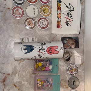 May include: A collection of Stray Kids merchandise, including round pins with character designs, a white pencil case with the Stray Kids logo, a tumbler, and small bags of decorative items. Also included are a photo card and keychains.