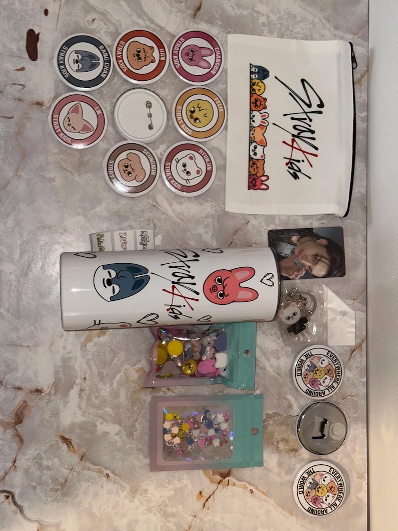 May include: A collection of Stray Kids merchandise, including round pins with character designs, a white pencil case with the Stray Kids logo, a tumbler, and small bags of decorative items. Also included are a photo card and keychains.