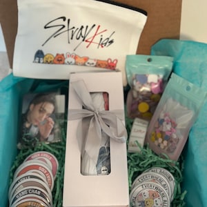 May include: An open gift box filled with merchandise. Includes a white zippered pouch with "Stray Kids" and cartoon characters, stickers, a photo, and a wrapped item. The box is lined with blue tissue paper and green shredded paper.