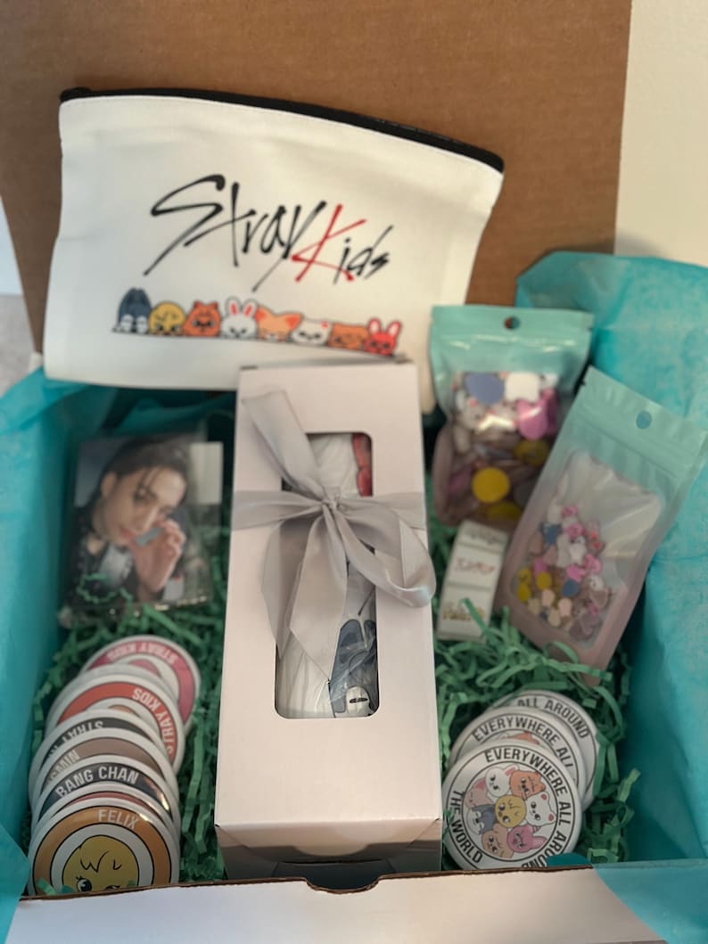May include: An open gift box filled with merchandise. Includes a white zippered pouch with "Stray Kids" and cartoon characters, stickers, a photo, and a wrapped item. The box is lined with blue tissue paper and green shredded paper.