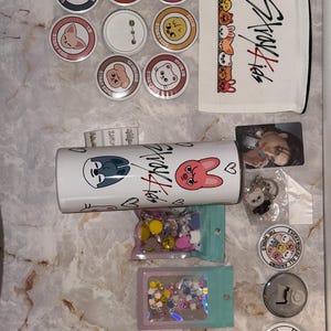 May include: A collection of Stray Kids merchandise, including buttons, a white tumbler, a zippered pouch, a photo card, and a keychain. The buttons feature cartoon character designs and the text "Stray Kids". The tumbler has the Stray Kids logo and cartoon characters.