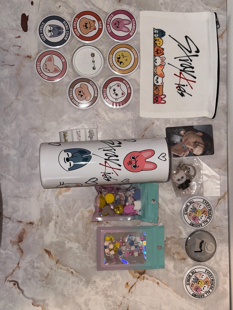 May include: A collection of Stray Kids merchandise, including buttons, a white tumbler, a zippered pouch, a photo card, and a keychain. The buttons feature cartoon character designs and the text "Stray Kids". The tumbler has the Stray Kids logo and cartoon characters.