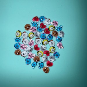 May include: A collection of colorful, cartoon-style character charms in various shapes, including hearts, bears, and round faces. The charms are in shades of red, blue, brown, yellow, and pink, set against a teal background.