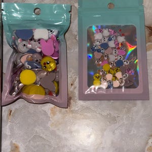 May include: Two sealed plastic bags filled with colorful, cartoon-shaped embellishments. The bags have a light blue top and a pink or light blue border. The embellishments include various animal shapes in shades of pink, yellow, gray, and blue.