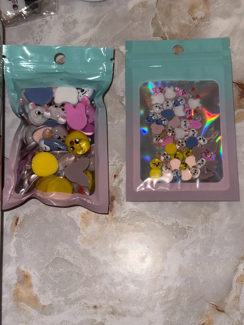 May include: Two sealed plastic bags filled with colorful, cartoon-shaped embellishments. The bags have a light blue top and a pink or light blue border. The embellishments include various animal shapes in shades of pink, yellow, gray, and blue.