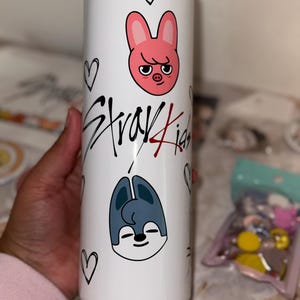 May include: A white tumbler featuring cartoon illustrations of a pink rabbit and a blue wolf, along with the text "Stray Kids" in black and red. The design includes black heart doodles. The tumbler is cylindrical and appears to be made of stainless steel.