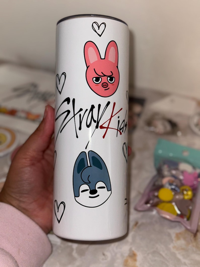 May include: A white tumbler featuring cartoon illustrations of a pink rabbit and a blue wolf, along with the text "Stray Kids" in black and red. The design includes black heart doodles. The tumbler is cylindrical and appears to be made of stainless steel.