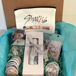 May include: A gift box filled with merchandise, including a white zippered pouch with "Stray Kids" text and cartoon characters, stickers, a photo card, a button, and other items. The box is lined with blue tissue paper and green shredded paper.