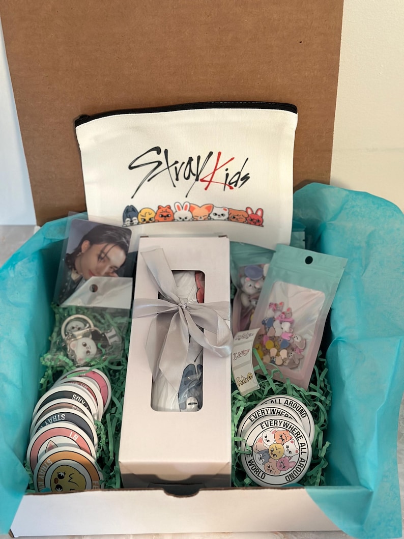 May include: A gift box filled with merchandise, including a white zippered pouch with "Stray Kids" text and cartoon characters, stickers, a photo card, a button, and other items. The box is lined with blue tissue paper and green shredded paper.