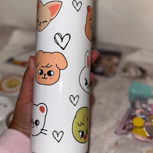 May include: White tumbler featuring cartoon animal faces and black heart doodles. The tumbler has a black lid and is cylindrical in shape. The animal faces include a fox, dog, bear, bunny, chick, and cat. The tumbler is designed for cold beverages.