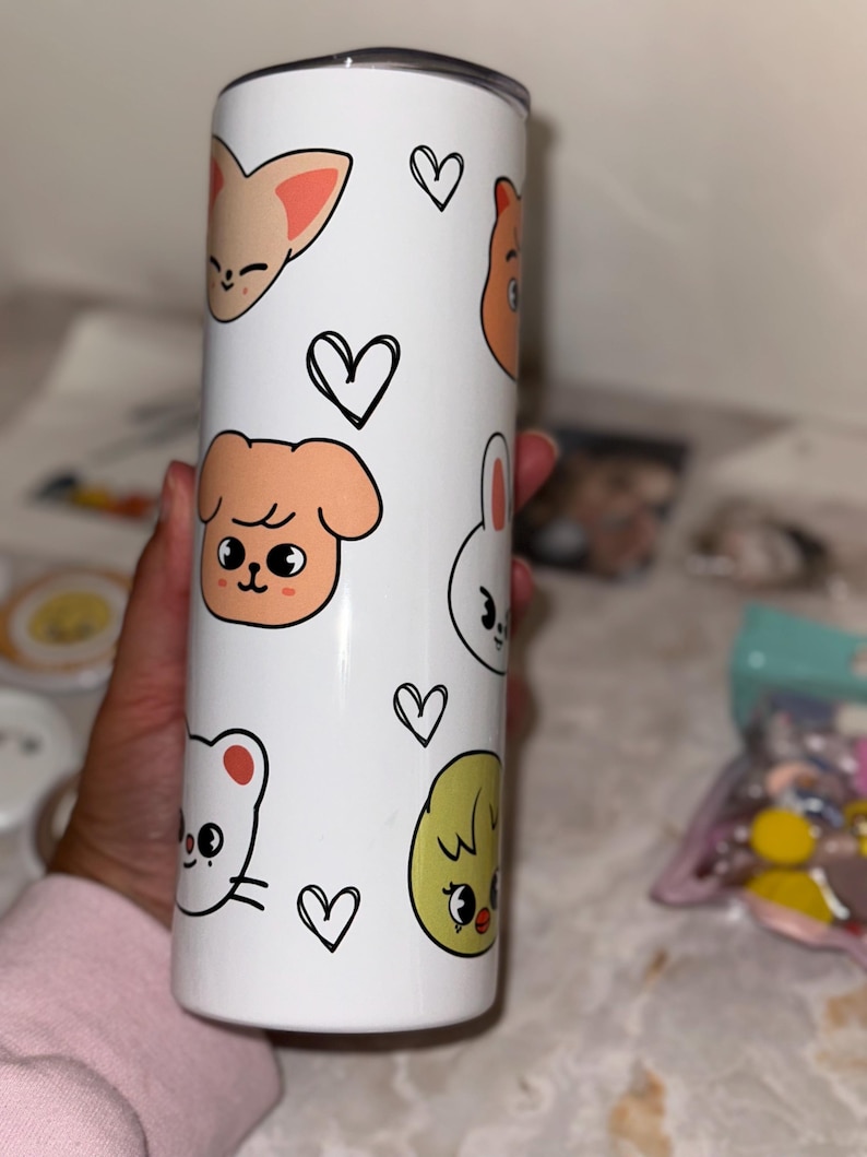 May include: White tumbler featuring cartoon animal faces and black heart doodles. The tumbler has a black lid and is cylindrical in shape. The animal faces include a fox, dog, bear, bunny, chick, and cat. The tumbler is designed for cold beverages.
