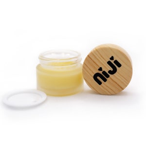 Peut inclure: Un petit pot en verre contenant un baume jaune. Le pot a un couvercle blanc et un couvercle en bois avec le mot "niji" imprimé dessus.