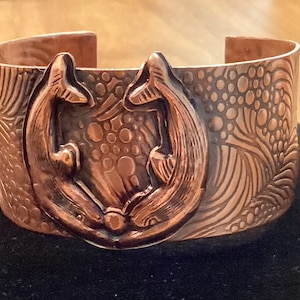 May include: A copper bracelet with a detailed design of two fish facing each other. The bracelet has a textured surface with a pattern of waves and bubbles.