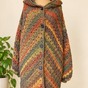 Könnte beinhalten: Eine Strickjacke mit Kapuze und Granny-Square-Häkelmuster in variierenden Erdtönen wie Olivgrün, gebranntem Orange, Senfgelb und Braun. Das Kleidungsstück verfügt über eine Knopfleiste an der Vorderseite mit vier dunklen, runden Knöpfen.