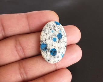 May include: Oval-shaped K2 stone with a white base, speckled with black and vibrant blue spots. The polished gemstone is held in a hand, showcasing its smooth surface and natural patterns. This unique stone is a beautiful addition to any jewelry collection.
