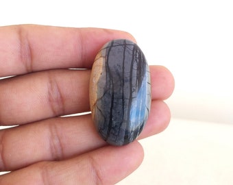 Puede incluir: Una gema ovalada con una superficie pulida. La piedra presenta una mezcla de colores gris, negro y beige, con patrones lineales oscuros que recuerdan a un paisaje. La gema se sostiene en una mano.