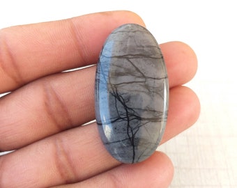 Puede incluir: Una piedra preciosa ovalada y pulida con un color base gris y azul. La piedra presenta intrincados patrones de vetas negras en toda su superficie, creando un diseño único y natural. La piedra preciosa se sostiene en una mano.