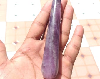 May include: A polished, elongated amethyst crystal wand. The wand is a deep purple color with lighter streaks and is held in an open hand. The background is a tiled surface.