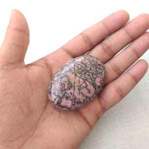 May include: An oval-shaped, polished gemstone with a pink base and intricate black veining. The stone is held in an open hand, showcasing its smooth texture and natural patterns. The gemstone's size is approximately 2 inches in length.